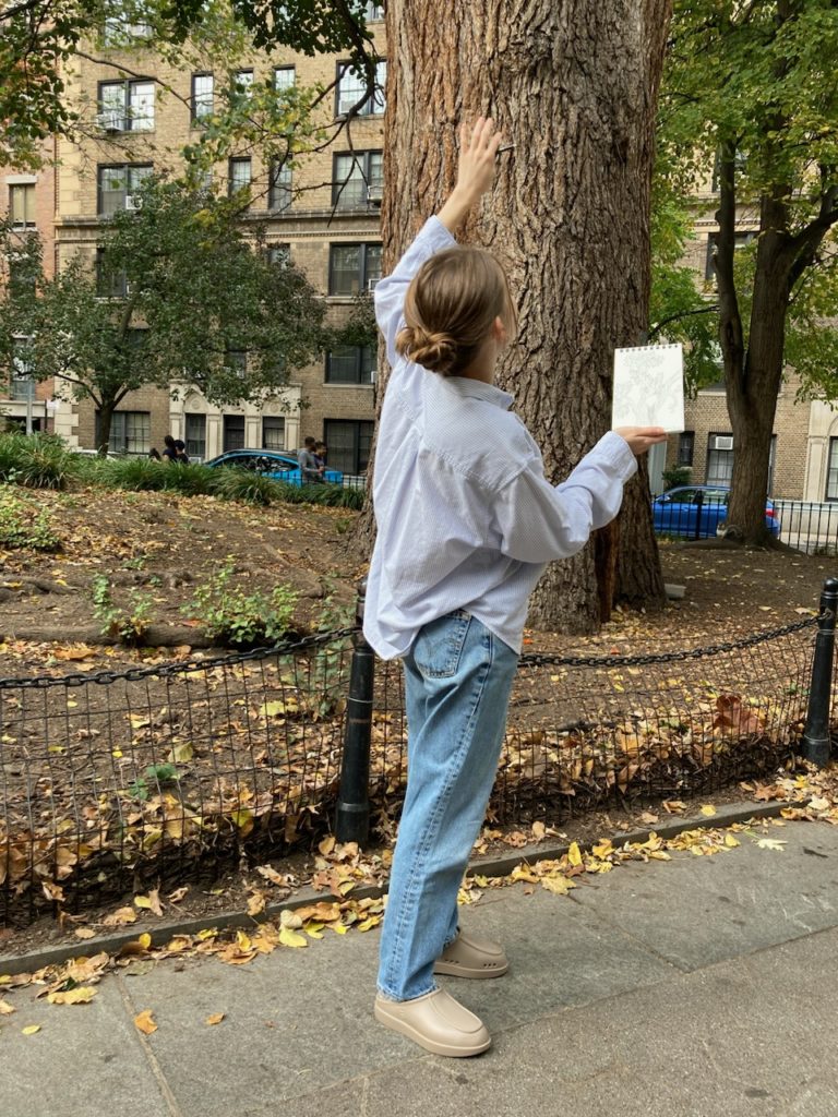 A woman with fair skin her back to the camera looking up and pointing to a large tree with one hand and there is a sketchbook in the other hand.