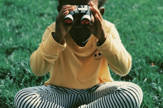 a child with dark skin wearing a yellow top and striped is holding binoculars up to her eyes while sitting cross legged on a lawn. Image via unsplash at photo-1540226410-cbd678d1e303.jpeg.