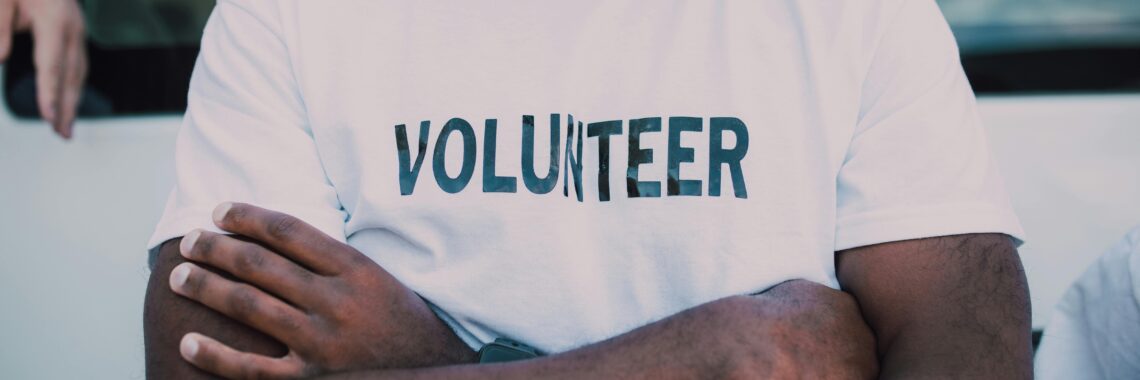 person with dark skin wearing a white short with the word "volunteer" in all caps on the front, crossing their arms across their chest. Photo by RDNE Stock project.